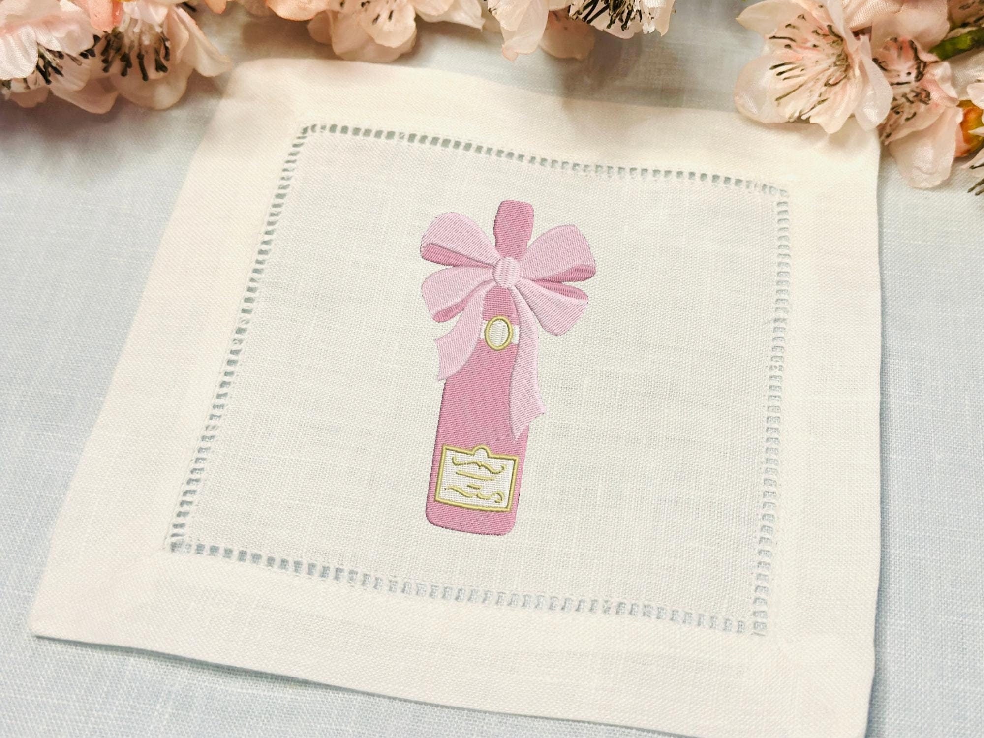 Beautifully embroidered pink champagne bottle with bow stitched onto a white linen cocktail napkin set on a light blue linen tablecloth with cherry blossoms at the top.