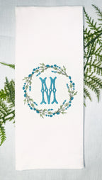 Custom personalized embroidered tea towel set on a light blue linen tablecloth and ferns. The white towel is embroidered with an initial (M) and surrounded in a wreath. The color of the embroidered initial and flowers in the wreath are teal.