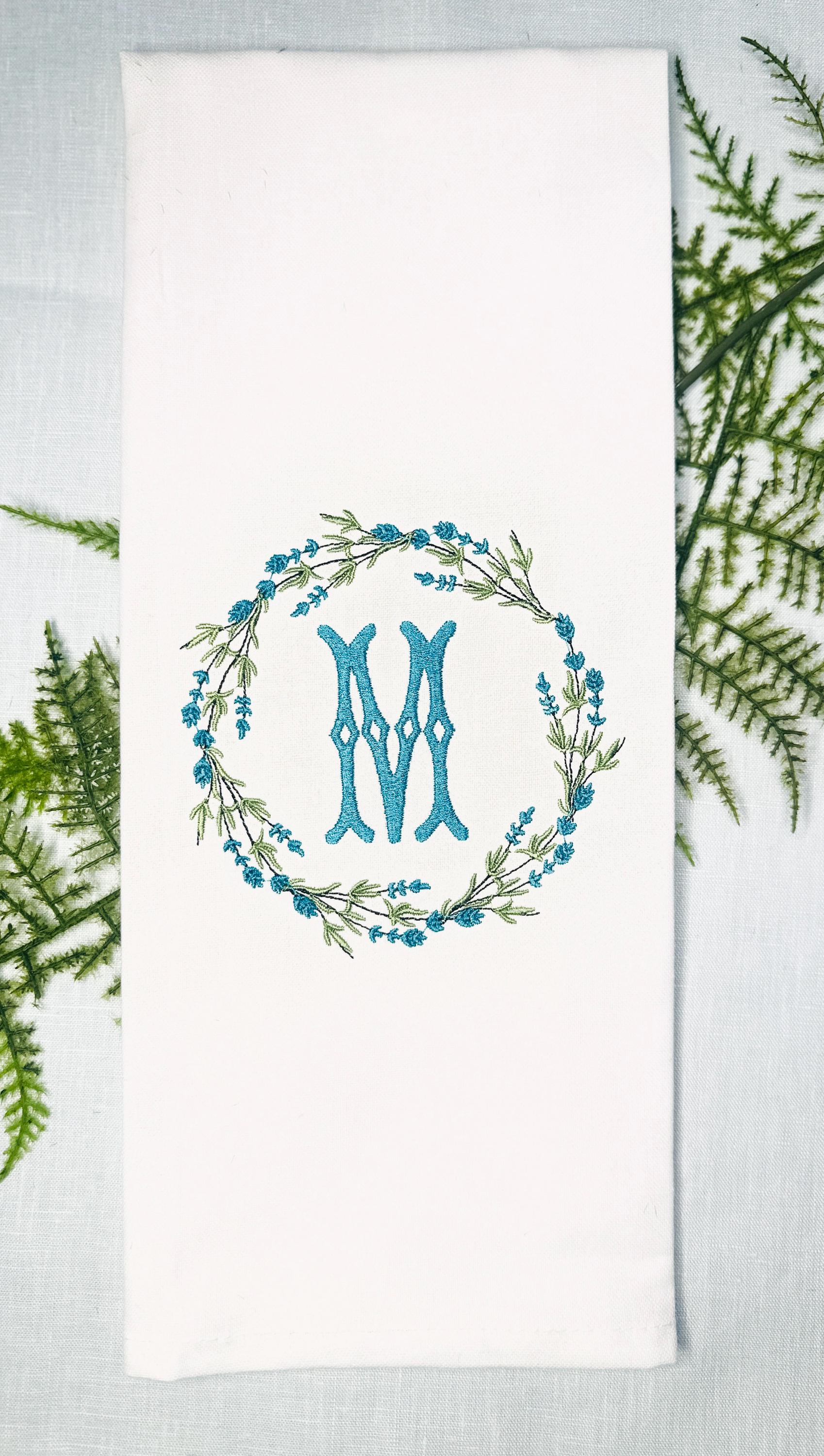 Custom personalized embroidered tea towel set on a light blue linen tablecloth and ferns. The white towel is embroidered with an initial (M) and surrounded in a wreath. The color of the embroidered initial and flowers in the wreath are teal.
