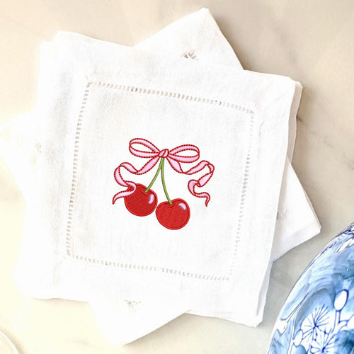 Cherries tied with a pink bow embroidered onto a white linen cocktail napkin. The napkins are in a pile on a white marble counter next to a blue and white vase. They are sized 6" square and have a hemstitch around the inside border.