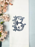 A personalized custom white tea towel with a navy blue letter B in Roman Font set on a linen baby blue tablecloth with cherry blossoms.