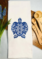A blue sea turtle ocean themed gift embroidered on a white tea towel set on a wooden cutting board with flowers and measuring spoons.