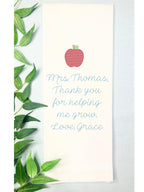 Teacher appreciation gift embroidered on a tea towel with an apple. Set on a blue linen table cloth with greens to the left.