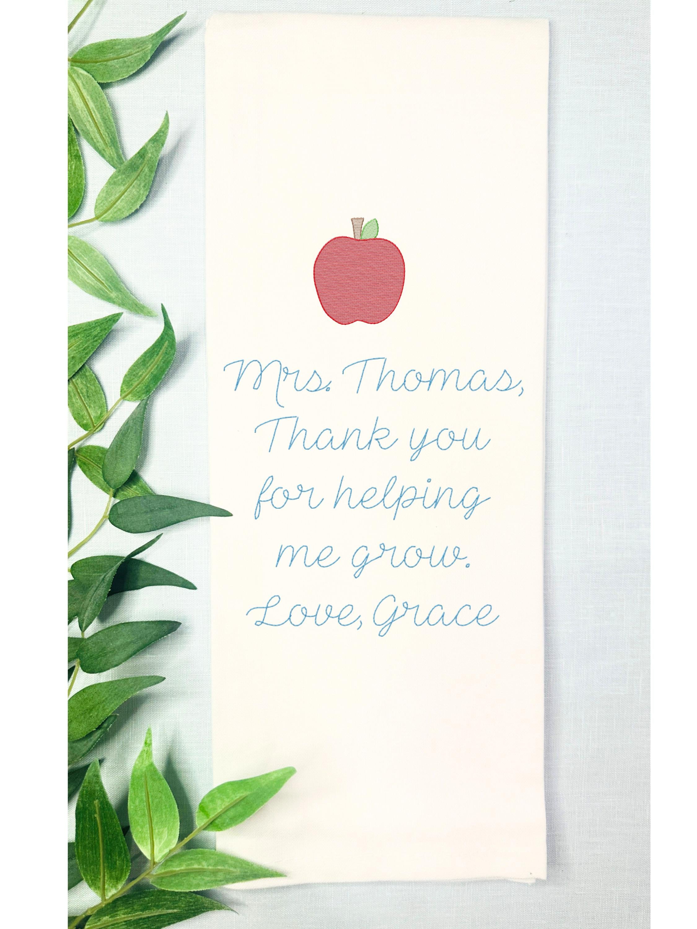 Teacher appreciation gift embroidered on a tea towel with an apple. Set on a blue linen table cloth with greens to the left.