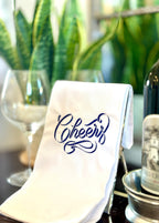 Cheers stitched in blue and white on a white tea towel for a 21st birthday gift for your sister! Set on a sidebar next to wine and wine glasses and a plant in the background.