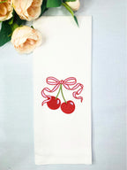 A white tea towel set on a blue linen background with red cherries and a pink and red bow embroidered on it. There are pale pink roses to the upper left.