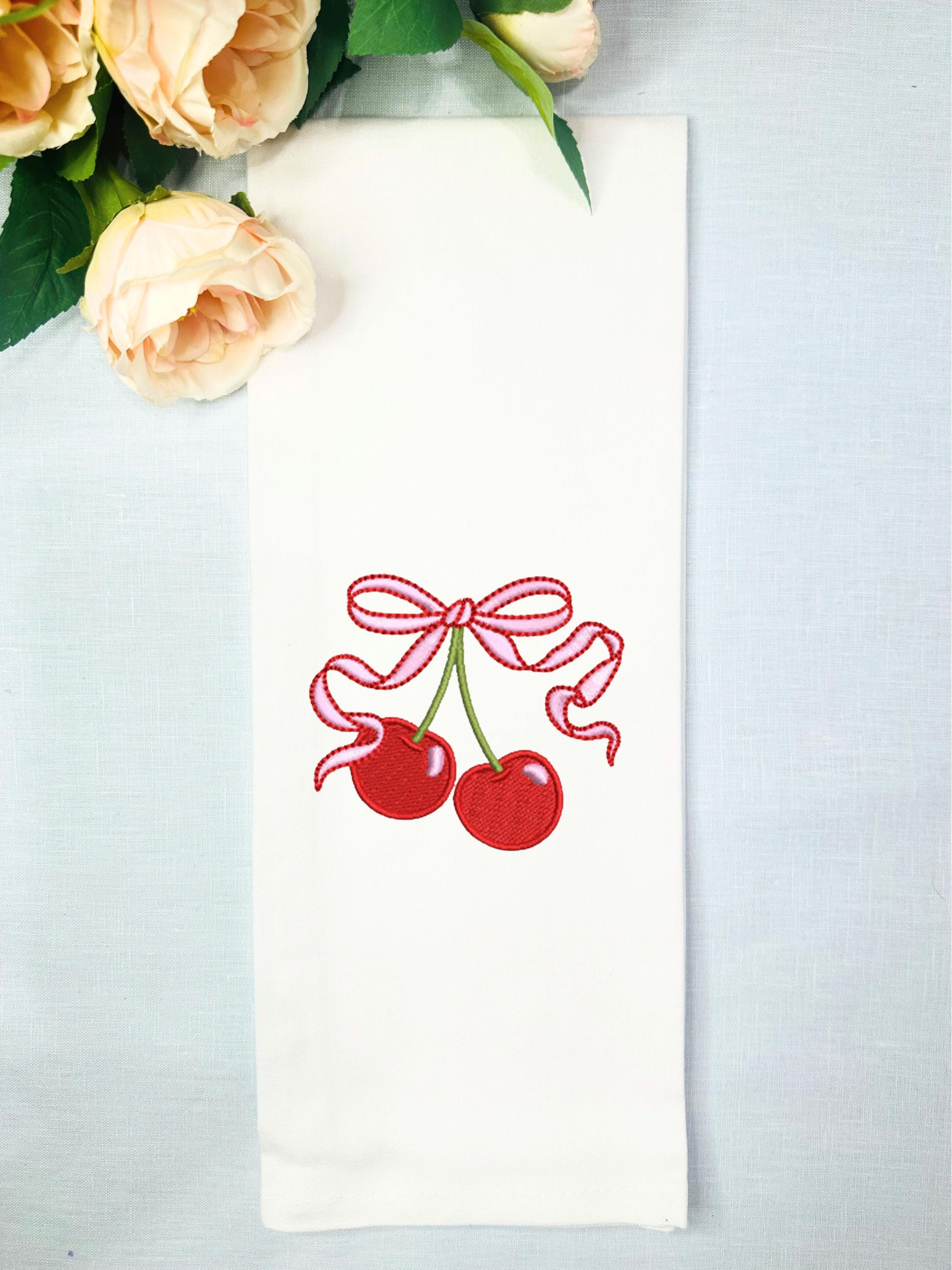 A white tea towel set on a blue linen background with red cherries and a pink and red bow embroidered on it. There are pale pink roses to the upper left.