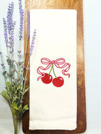 Cherry Bar Cart tea towel. Red Cherries with pink bow set on a cutting board on top of a haint blue linen tablecloth with lavender to the left side.