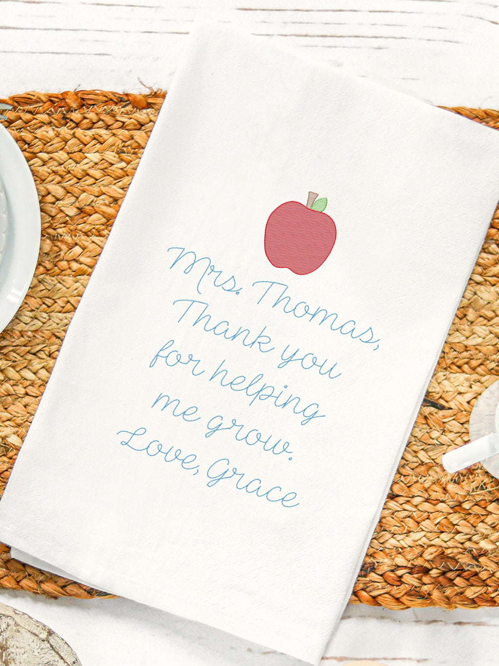 Teacher appreciation gift of an embroidered tea towel with a message that says: Mrs. Thomas, Thank you for helping me grow. Love, Grace. There is an apple on top of the message, also embroidered. It is set on a jute placemat with coffee cup & saucer.