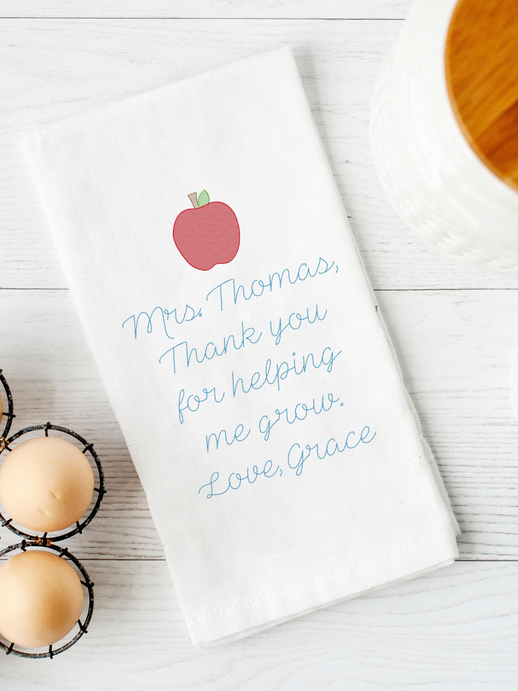 A white kitchen towel embroidered with a red apple and blue text that reads &#39;Mrs. Thomas, Thank you for helping me grow. Love, Grace.&#39; The towel is set on a white board table next to eggs and a white canister with a wooden top.