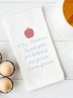 A white kitchen towel embroidered with a red apple and blue text that reads &#39;Mrs. Thomas, Thank you for helping me grow. Love, Grace.&#39; The towel is set on a white board table next to eggs and a white canister with a wooden top.