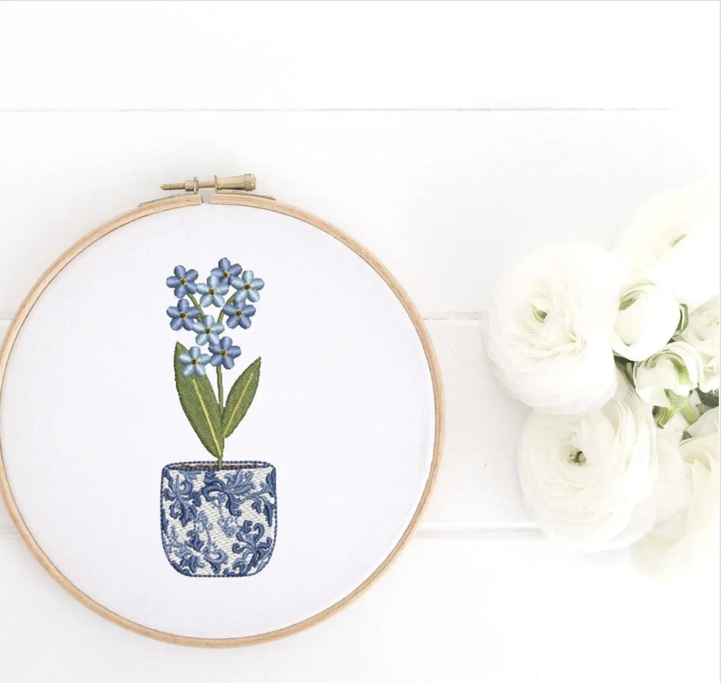 Different tones of soft blue flowers in a chinoiserie style pot embroidered and set in a wooden hoop. It is set on a white wooden background next to white roses.