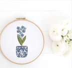 Different tones of soft blue flowers in a chinoiserie style pot embroidered and set in a wooden hoop. It is set on a white wooden background next to white roses.