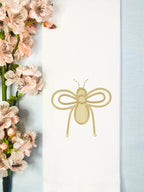 A kitchen towel with an embroidered bee for home decor set on a linen baby blue tablecloth with cherry blossoms to the left.