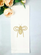 A spring bee decor tea towel that has a honeybee embroidered in gold on a white tea towel set on a table with a light blue linen background with peach roses.