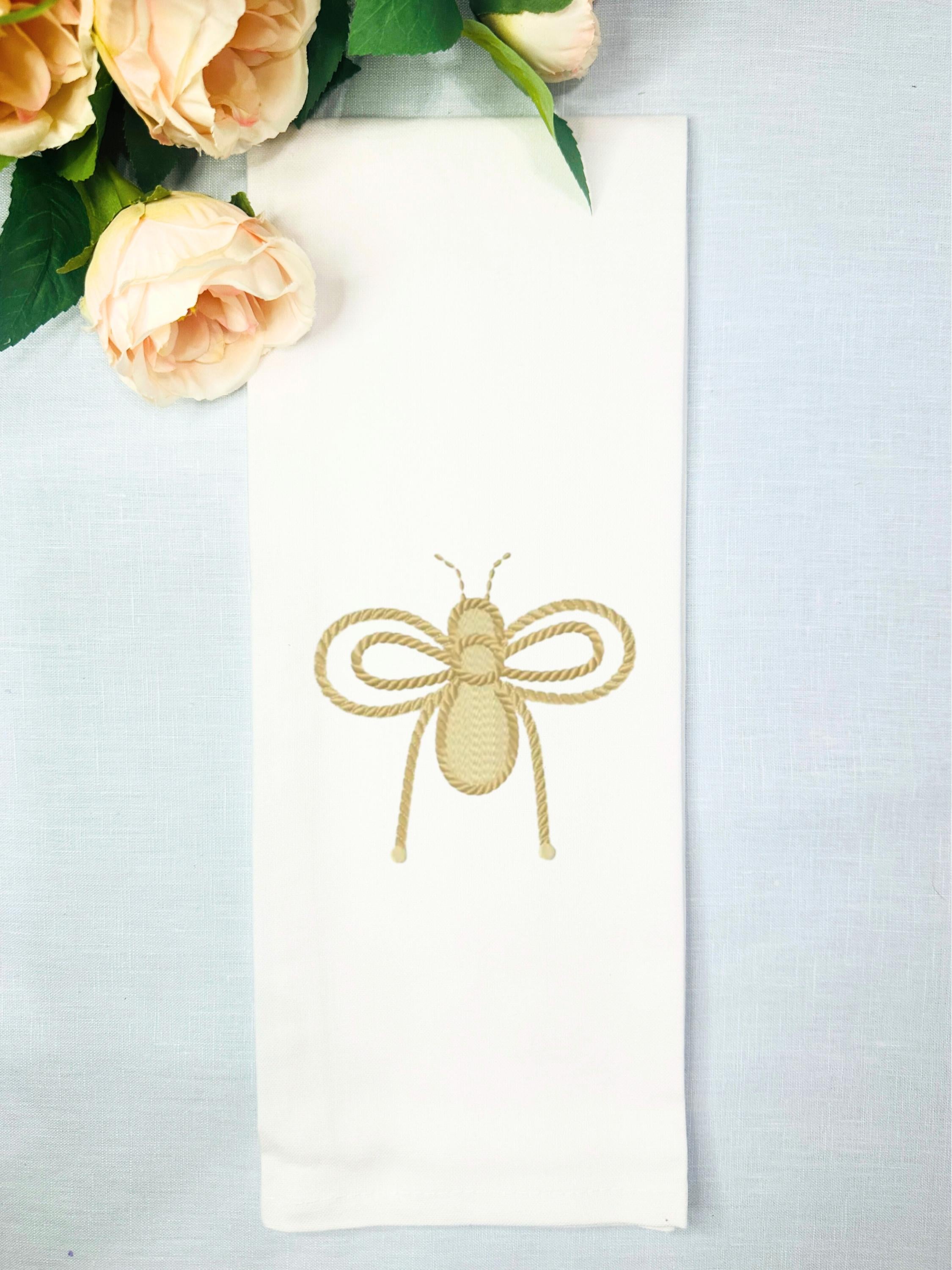 A spring bee decor tea towel that has a honeybee embroidered in gold on a white tea towel set on a table with a light blue linen background with peach roses.