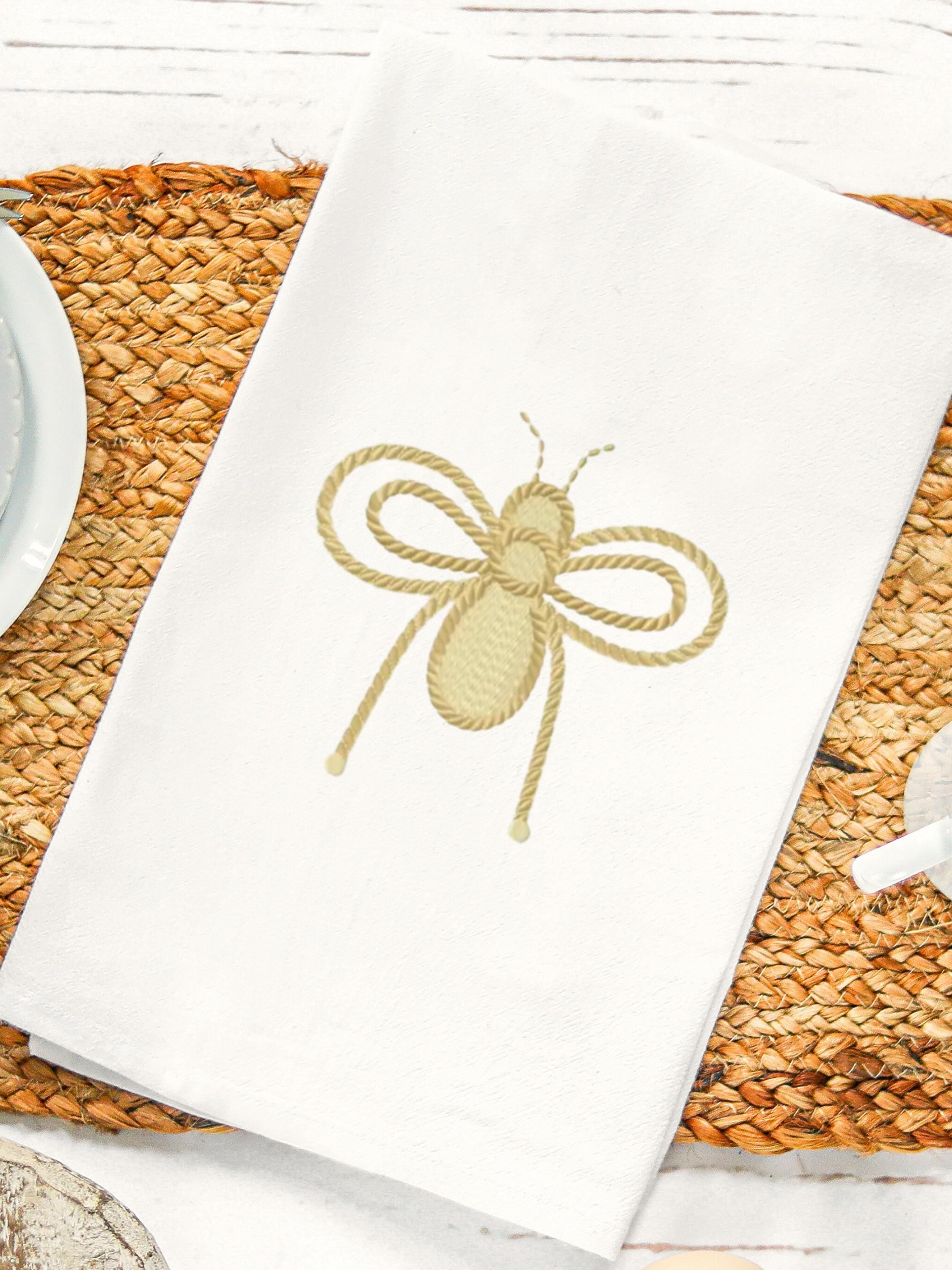 A white 100% cotton tea towel with hanging loop is embroidered with a gold queen bee and set on a wicker placemat on a white table with a white saucer to the left and a white cup to the right. Tea towels measure approximately 17” wide by 30” long.