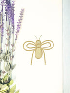 A honey bee embroidered in gold on a white tea towel on a light blue linen tablecloth with lavender next to it.