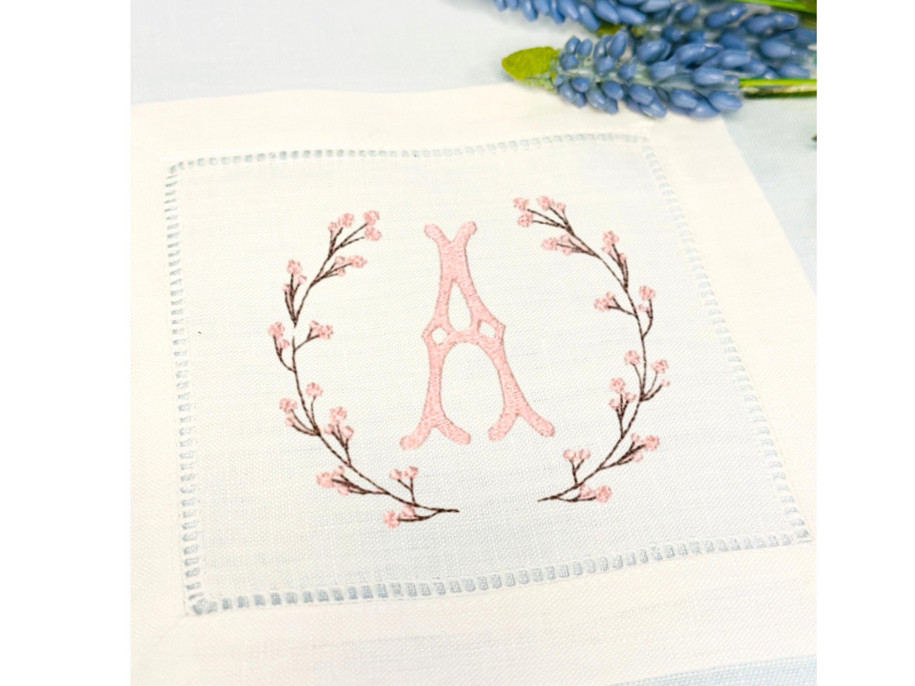 A set of 4 cocktail napkins embroidered with the letter A in a cherry blossom frame. The cherry blossoms are pink, the stems are brown, and the initial matches the pink of the cherry blossoms. They are set on a blue tablecloth with a blue flower stem to the upper right.