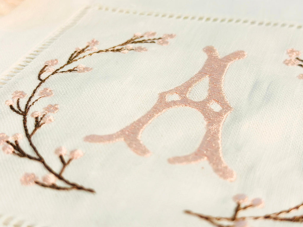 A close up of A set of 4 cocktail napkins embroidered with the letter A in a cherry blossom frame. The cherry blossoms are pink, the stems are brown, and the initial matches the pink of the cherry blossom