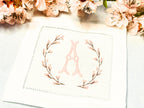 A set of 4 cocktail napkins embroidered with the letter A in a cherry blossom frame. The cherry blossoms are pink, the stems are brown, and the initial matches the pink of the cherry blossoms. They are set on a table with cherry blossoms near the top.