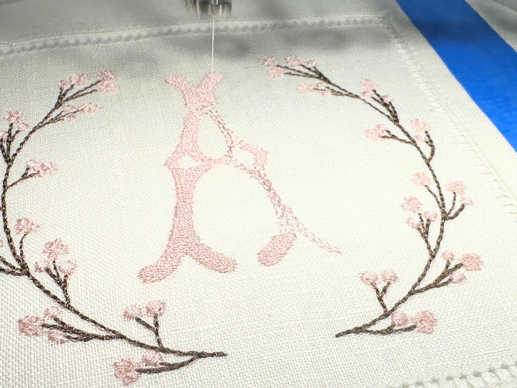 A set of 4 cocktail napkins embroidered with the letter A in a cherry blossom frame being embroidered. The cherry blossoms are pink, the stems are brown, and the initial matches the pink of the cherry blossoms.