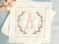 A set of 4 cocktail napkins embroidered with the letter A in a cherry blossom frame. The cherry blossoms are pink, the stems are brown, and the initial matches the pink of the cherry blossoms. They are set on a light blue linen background with faux cherry blossoms on the upper left of the photo.