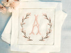 A set of 4 cocktail napkins embroidered with the letter A in a cherry blossom frame. The cherry blossoms are pink, the stems are brown, and the initial matches the pink of the cherry blossoms. They are set on a light blue linen background with faux cherry blossoms on the upper left of the photo.