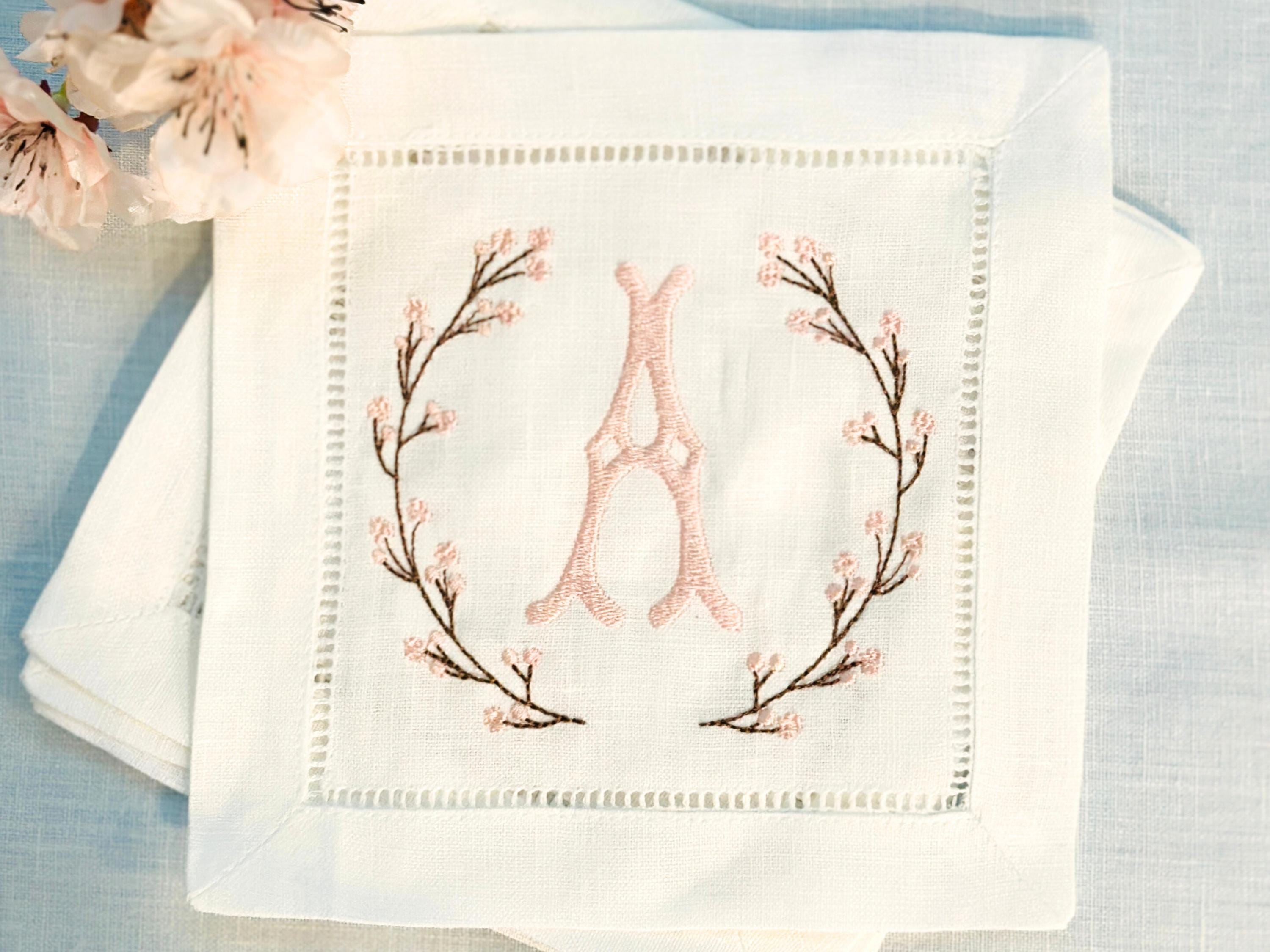 A set of 4 cocktail napkins embroidered with the letter A in a cherry blossom frame. The cherry blossoms are pink, the stems are brown, and the initial matches the pink of the cherry blossoms. They are set on a light blue linen background with faux cherry blossoms on the upper left of the photo.