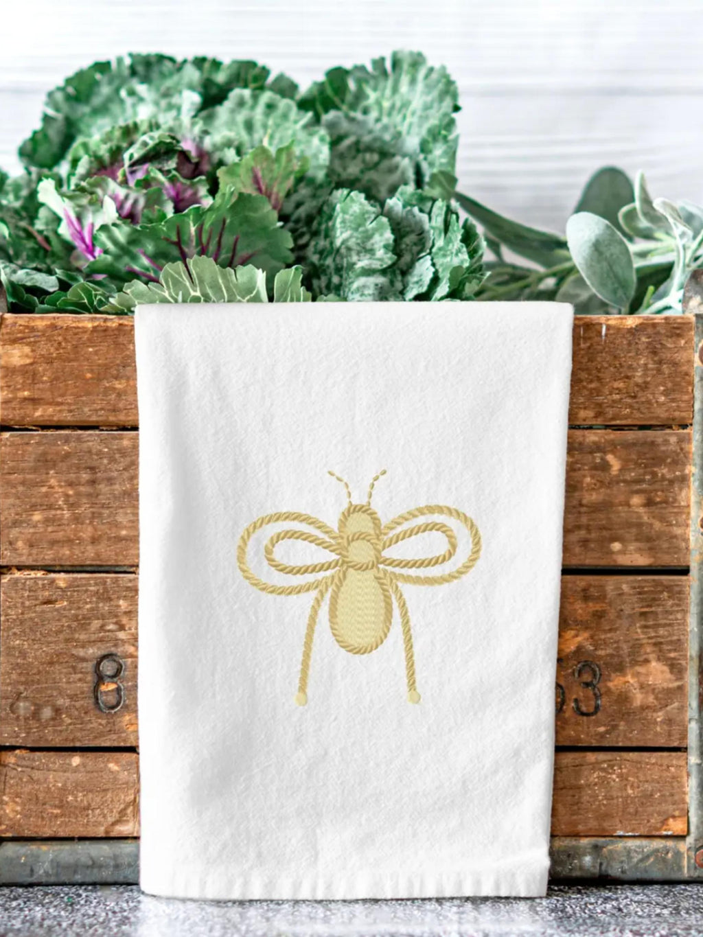 An embroidered gold bee on a white dish towel on the edge of a market bin holding cabbage.