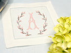 A set of 4 cocktail napkins embroidered with the letter A in a cherry blossom frame. The cherry blossoms are pink, the stems are brown, and the initial matches the pink of the cherry blossoms. They are set on a blue linen tablecloth with a white hydrangea on the bottom right.