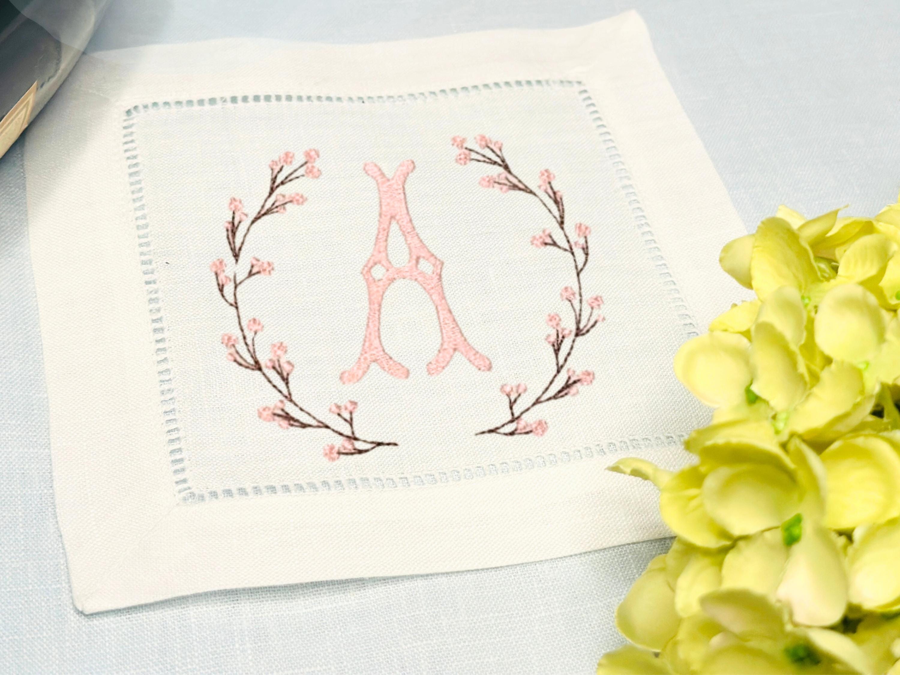 A set of 4 cocktail napkins embroidered with the letter A in a cherry blossom frame. The cherry blossoms are pink, the stems are brown, and the initial matches the pink of the cherry blossoms. They are set on a blue linen tablecloth with a white hydrangea on the bottom right.