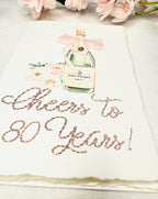 A birthday card with a watercolor champagne bottle with a pink bow and flowers to the left side. Under the art is an embroidered message: Cheers to 80 Years in pink thread. The card and matching envelope are set on a white linen tablecloth with pink cherry blossoms on top.