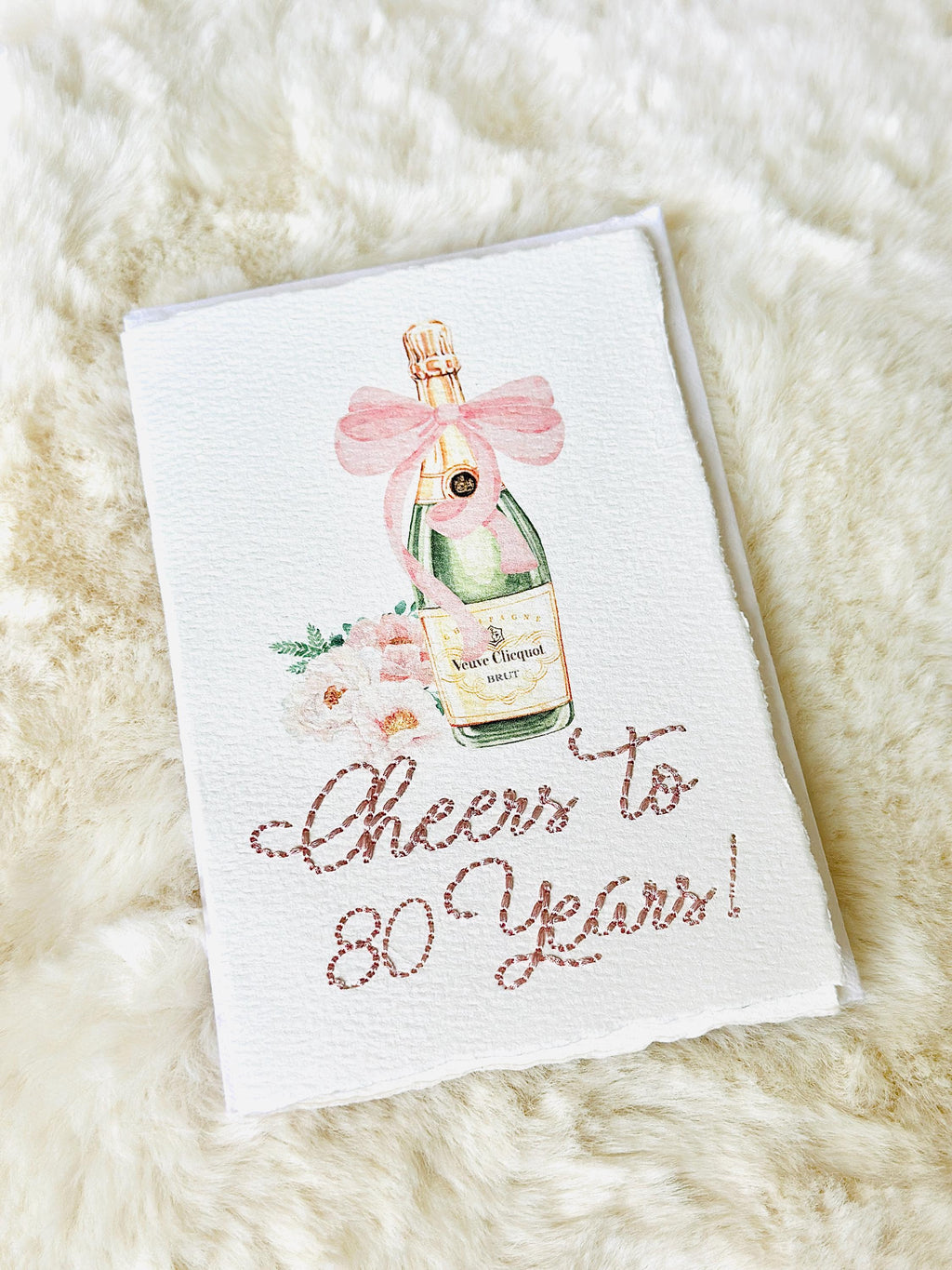A birthday card with a watercolor champagne bottle with a pink bow and flowers to the left side. Under the art is an embroidered message: Cheers to 80 Years in pink thread. The card and matching envelope are set on a fur blanket.