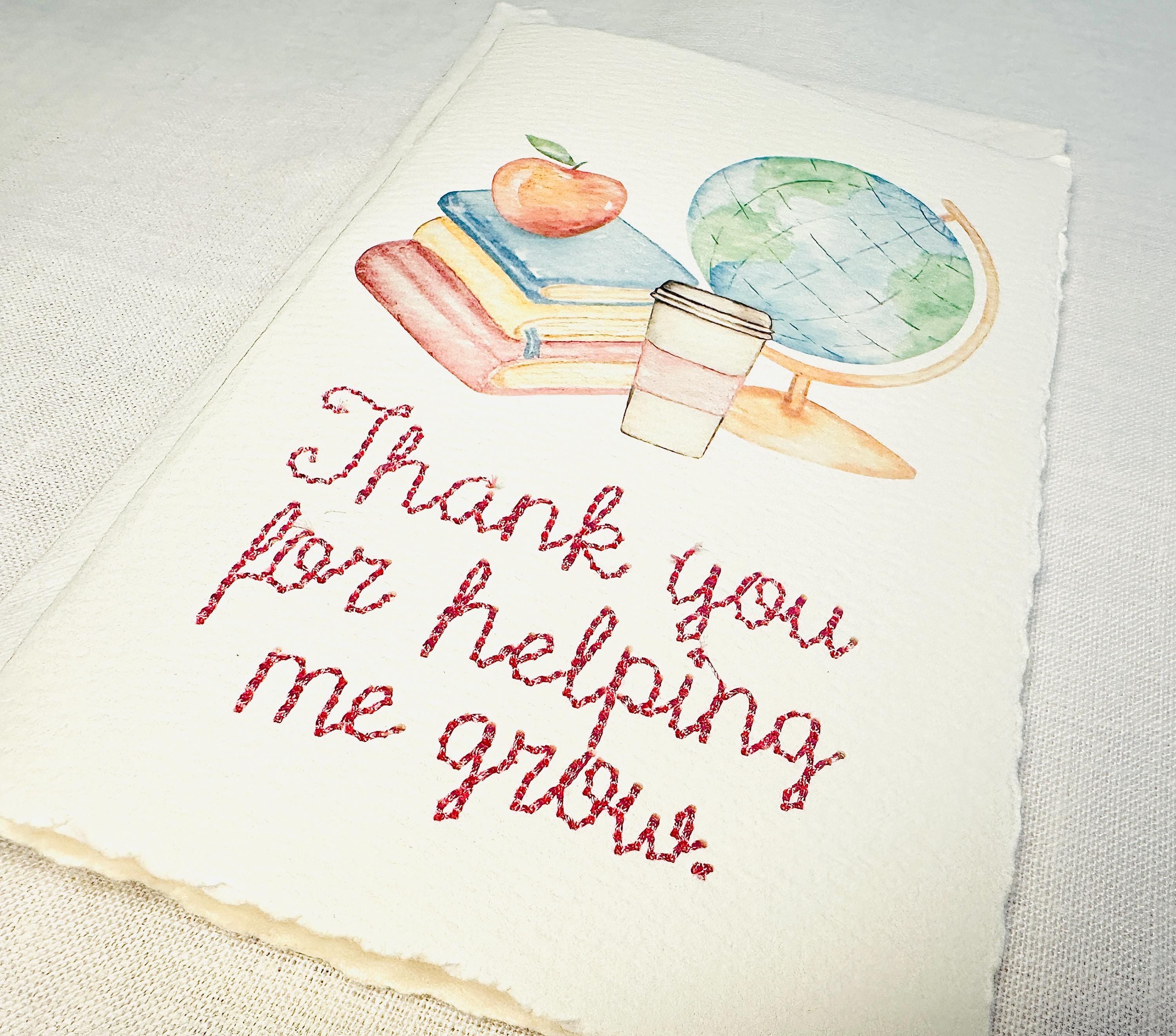 A handmade card that has watercolor art of a pile of books with an apple on top next to a globe and a to-go cup of coffee in soft pastel colors with embroidery under it that says: Thank you for helping me grow. The embroidery is done in pink thread. The card is set on a white table.