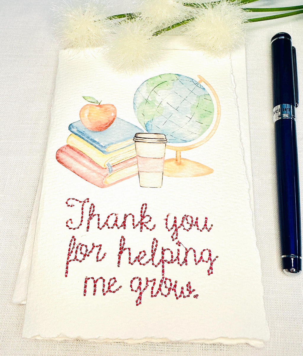 A handmade card that has watercolor art of a pile of books with an apple on top next to a globe and a to-go cup of coffee in soft pastel colors with embroidery under it that says: Thank you for helping me grow. The embroidery is done in pink thread. The card is set on a white table with a pen to the right and white puffy flowers on top.