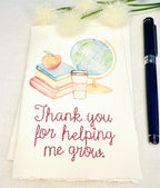 A handmade card that has watercolor art of a pile of books with an apple on top next to a globe and a to-go cup of coffee in soft pastel colors with embroidery under it that says: Thank you for helping me grow. The embroidery is done in pink thread. The card is set on a white table with a pen to the right and white puffy flowers on top.