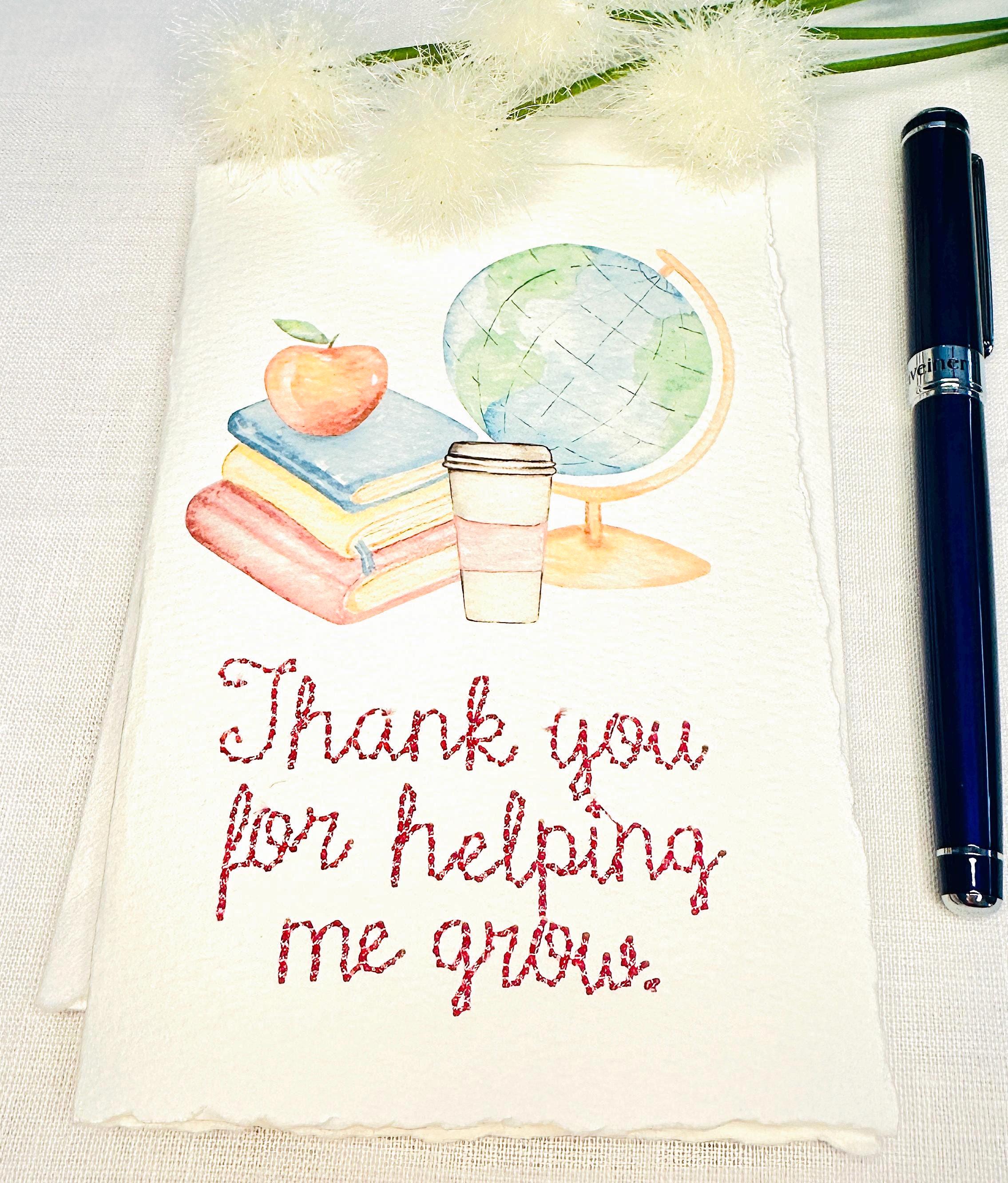 A handmade card that has watercolor art of a pile of books with an apple on top next to a globe and a to-go cup of coffee in soft pastel colors with embroidery under it that says: Thank you for helping me grow. The embroidery is done in pink thread. The card is set on a white table with a pen to the right and white puffy flowers on top.