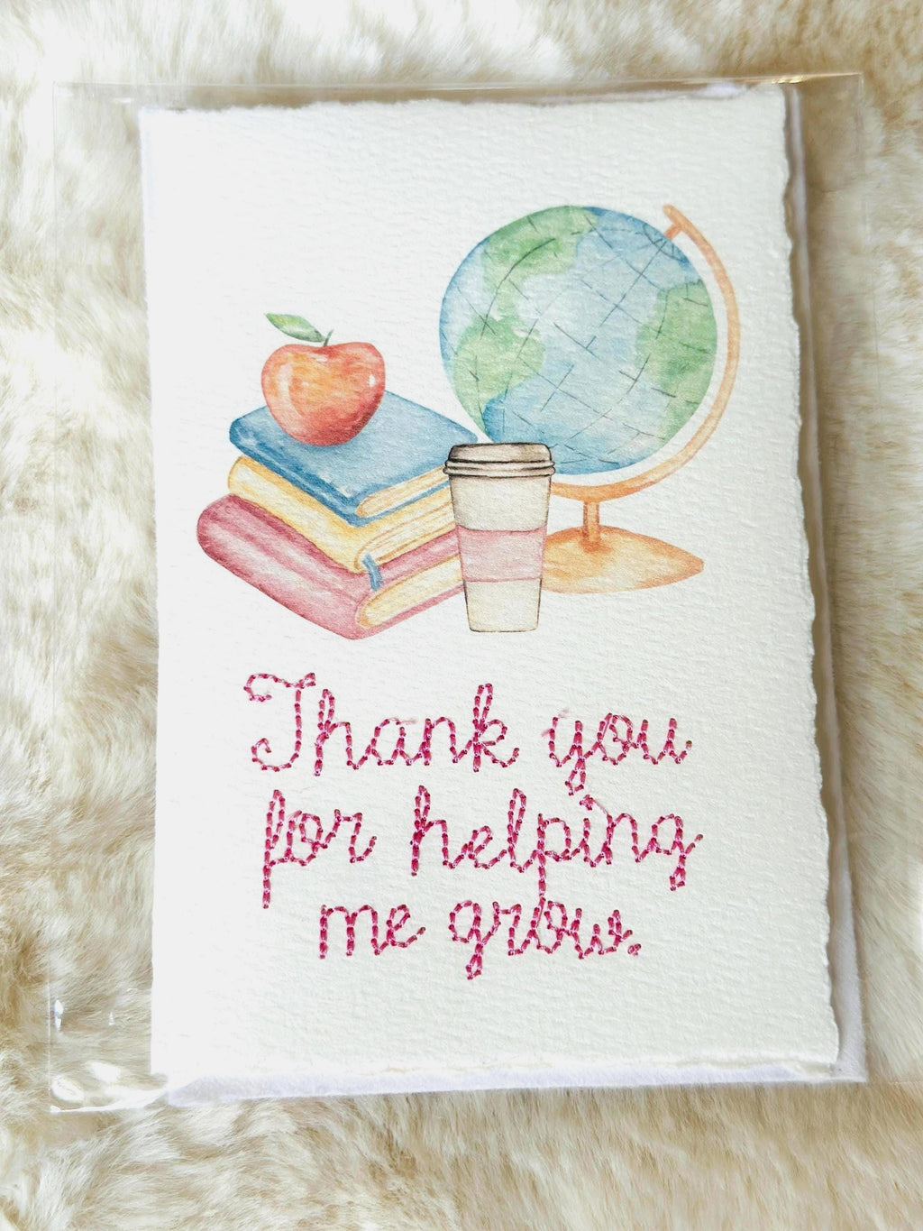 A handmade card that has watercolor art of a pile of books with an apple on top next to a globe and a to-go cup of coffee in soft pastel colors with embroidery under it that says: Thank you for helping me grow. The embroidery is done in pink thread. The card is set on a fur mat.