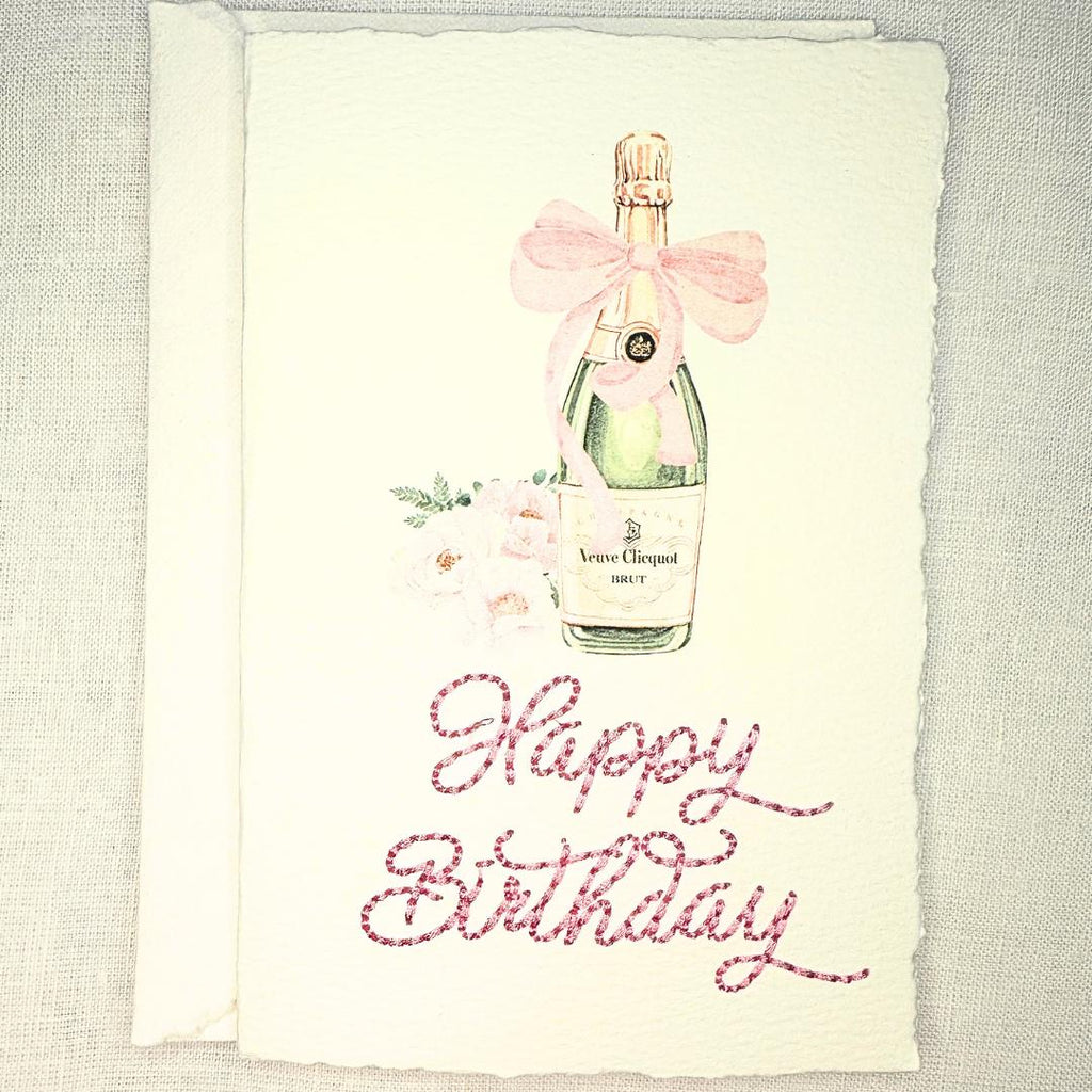A deckled edge card with watercolor art of a champagne bottle with a pink bow and pink flowers to the left. Under the art is embroidered: Happy Birthday in pink. The card is set on a white linen table. Cards are approximately 4x6.