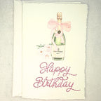 A deckled edge card with watercolor art of a champagne bottle with a pink bow and pink flowers to the left. Under the art is embroidered: Happy Birthday in pink. The card is set on a white linen table. Cards are approximately 4x6.