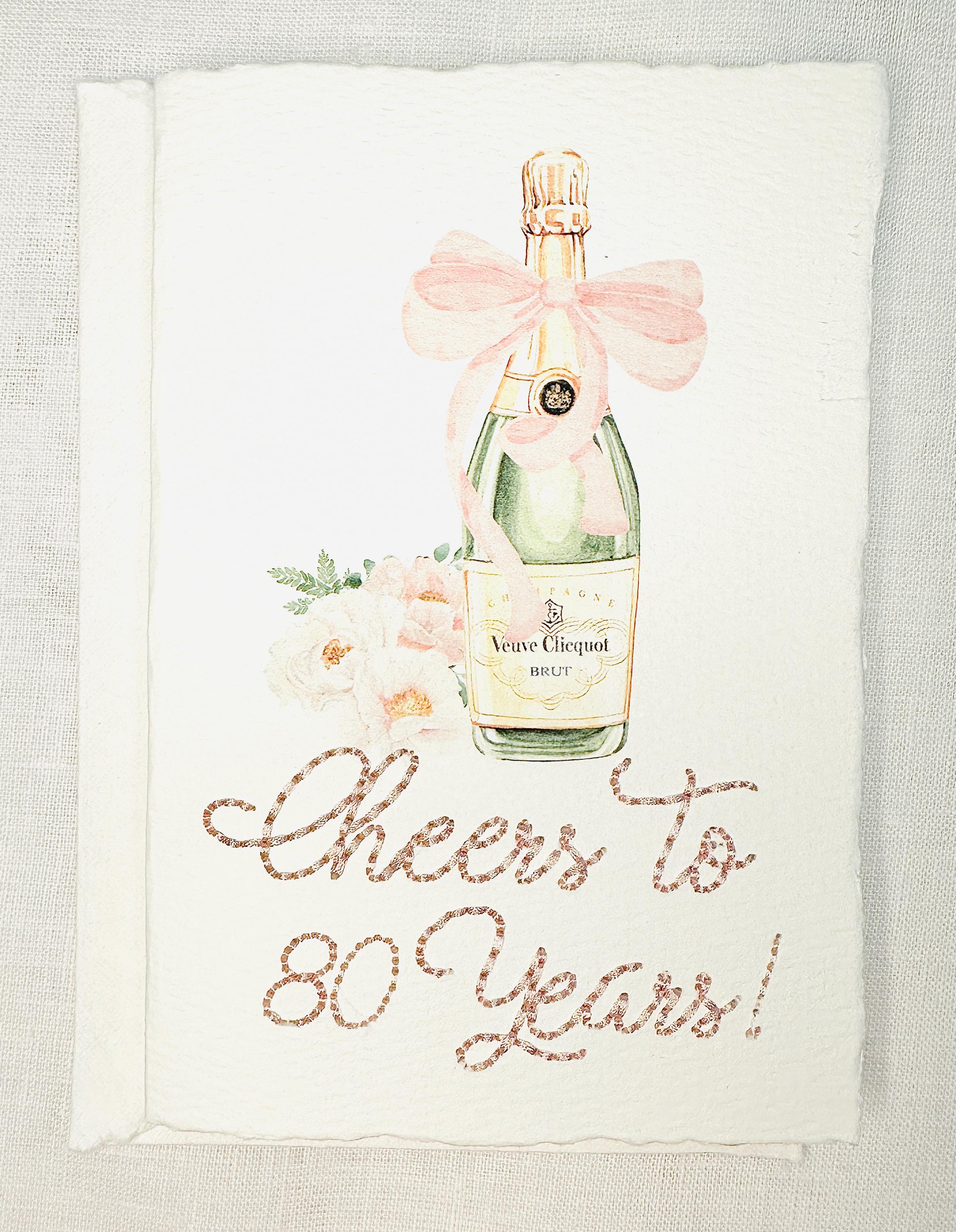 A birthday card with a watercolor champagne bottle with a pink bow and flowers to the left side. Under the art is an embroidered message: Cheers to 80 Years in pink thread. The card and matching envelope are set on a white linen tablecloth.