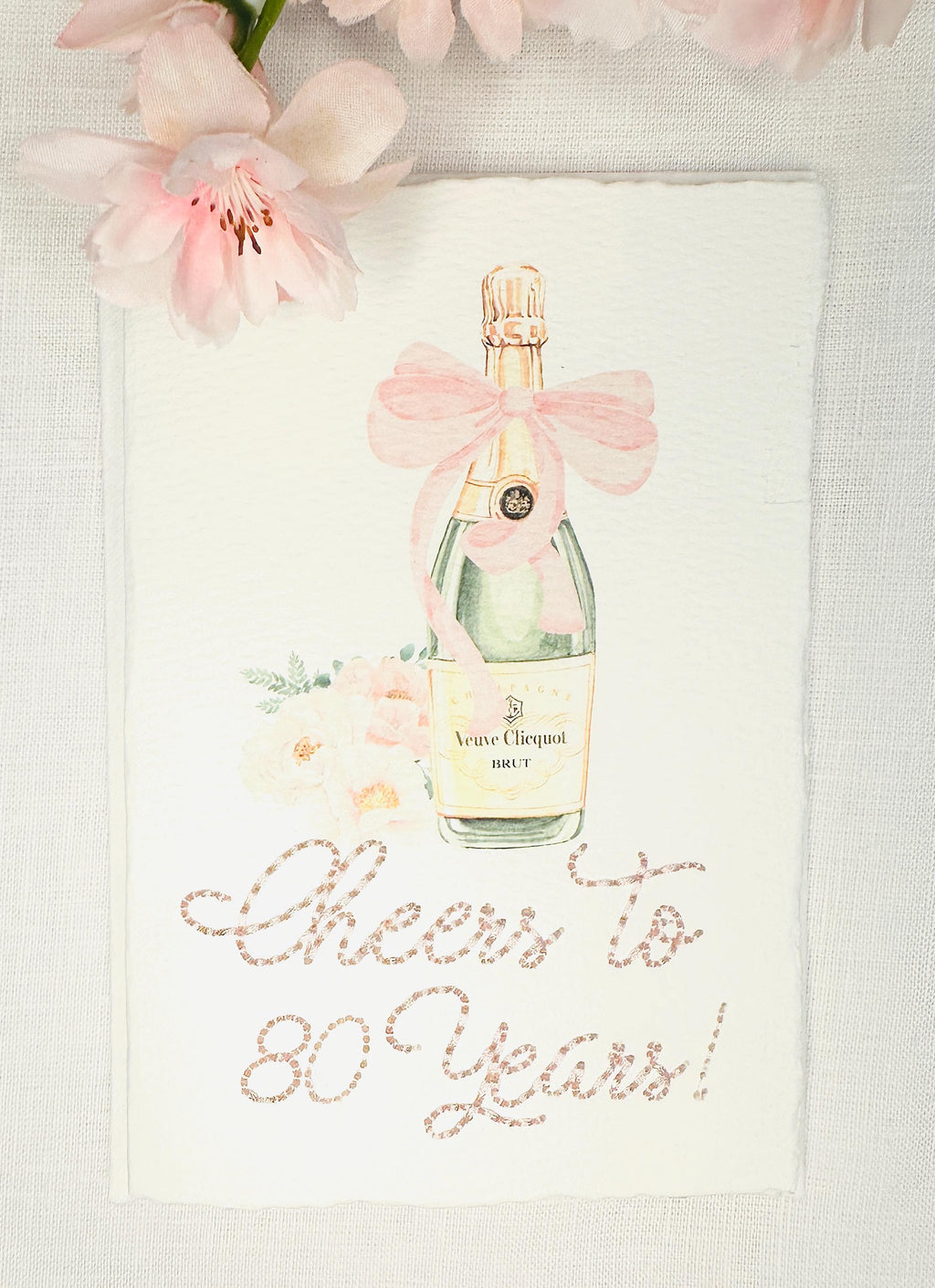 A birthday card with a watercolor champagne bottle with a pink bow and flowers to the left side. Under the art is an embroidered message: Cheers to 80 Years in pink thread. The card and matching envelope are set on a white linen tablecloth with pink flowers on the top of the card.
