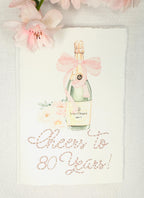 A birthday card with a watercolor champagne bottle with a pink bow and flowers to the left side. Under the art is an embroidered message: Cheers to 80 Years in pink thread. The card and matching envelope are set on a white linen tablecloth with pink flowers on the top of the card.