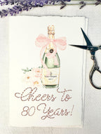 A birthday card with a watercolor champagne bottle with a pink bow and flowers to the left side. Under the art is an embroidered message: Cheers to 80 Years in pink thread. The card and matching envelope are set on a white linen tablecloth with a sprig on lavender over the top of the card and embroidery scissors to the right.