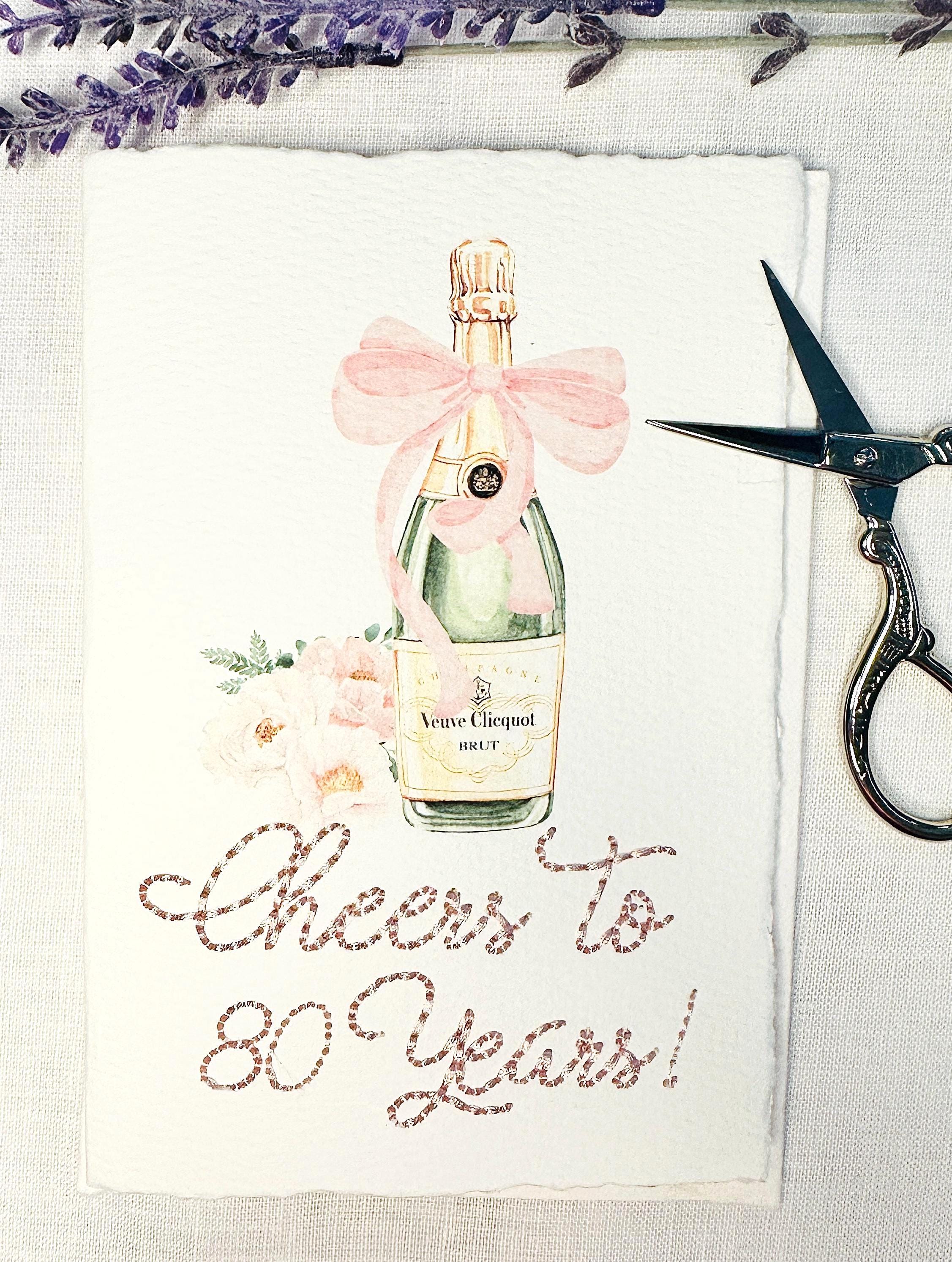 A birthday card with a watercolor champagne bottle with a pink bow and flowers to the left side. Under the art is an embroidered message: Cheers to 80 Years in pink thread. The card and matching envelope are set on a white linen tablecloth with a sprig on lavender over the top of the card and embroidery scissors to the right.