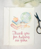 A handmade card that has watercolor art of a pile of books with an apple on top next to a globe and a to-go cup of coffee in soft pastel colors with embroidery under it that says: Thank you for helping me grow. The embroidery is done in pink thread. The card is set on its&#39; envelope on a white table with embroidery scissors to tthe right and white puffy stems on the top.