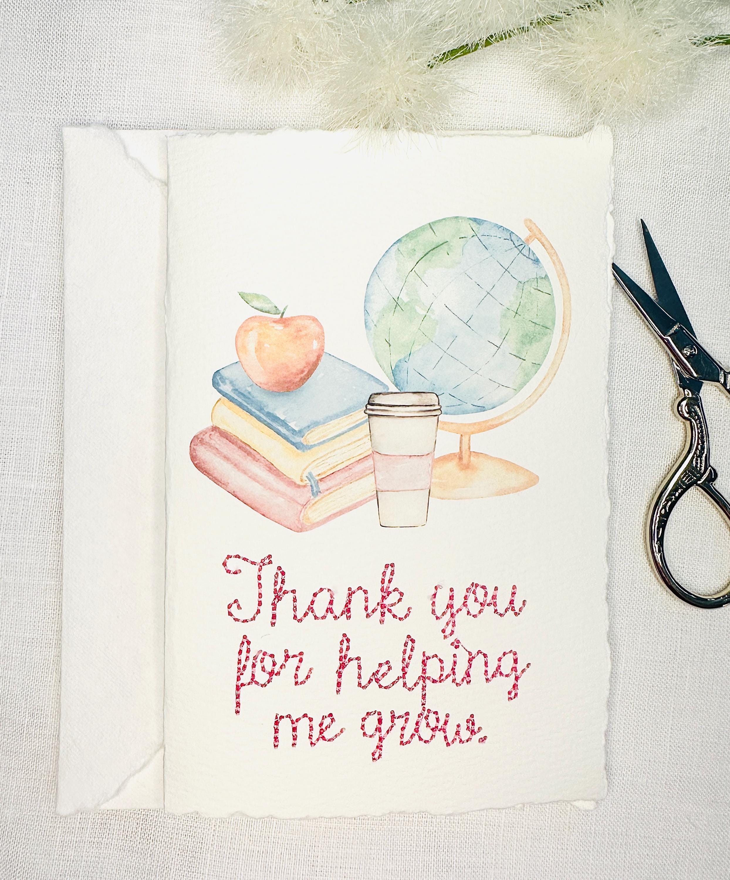 A handmade card that has watercolor art of a pile of books with an apple on top next to a globe and a to-go cup of coffee in soft pastel colors with embroidery under it that says: Thank you for helping me grow. The embroidery is done in pink thread. The card is set on its&#39; envelope on a white table with embroidery scissors to tthe right and white puffy stems on the top.
