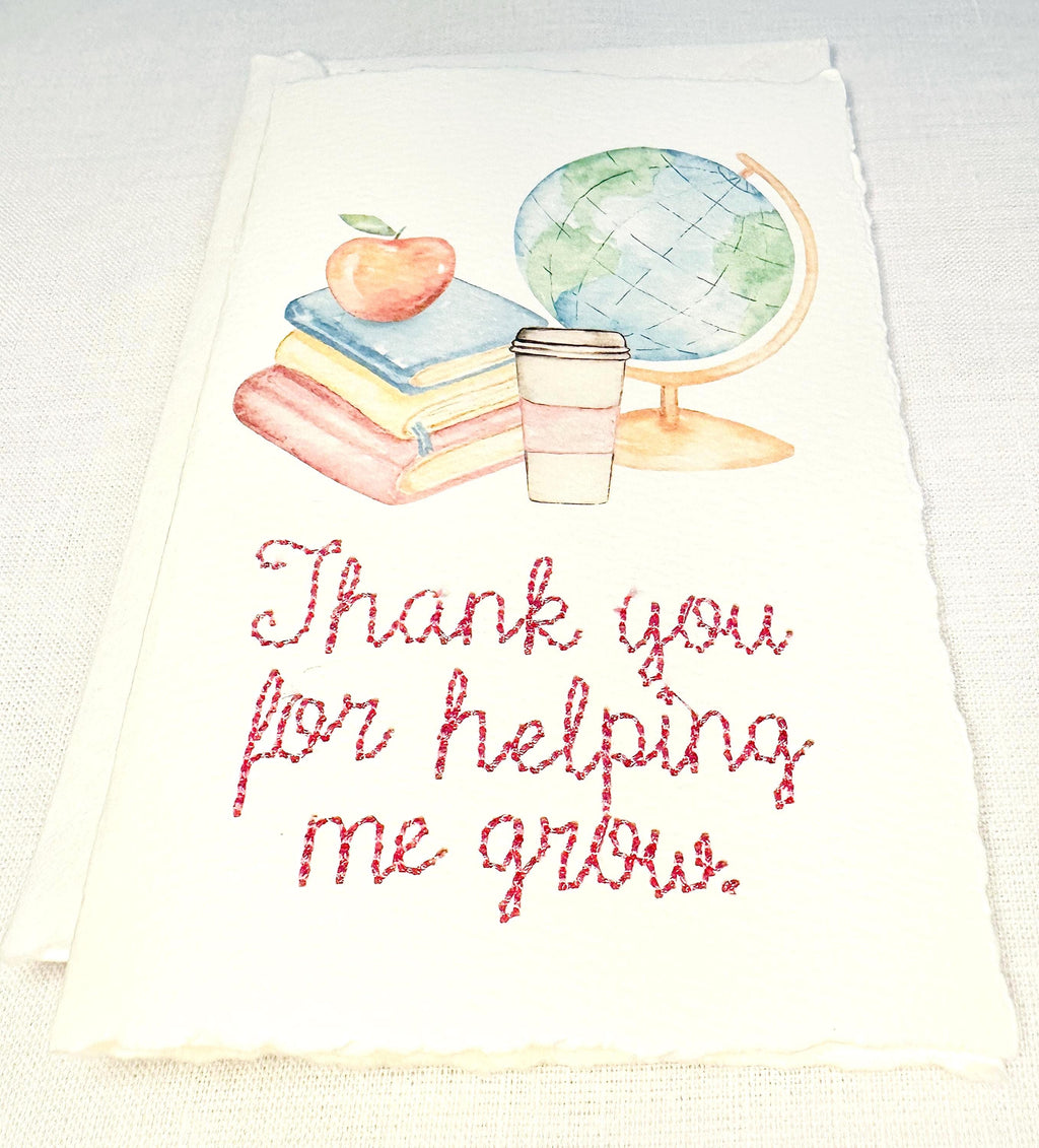 A handmade card that has watercolor art of a pile of books with an apple on top next to a globe and a to-go cup of coffee in soft pastel colors with embroidery under it that says: Thank you for helping me grow. The embroidery is done in pink thread. The card is set on a white table.