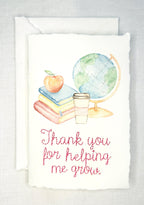 A handmade card that has watercolor art of a pile of books with an apple on top next to a globe and a to-go cup of coffee in soft pastel colors with embroidery under it that says: Thank you for helping me grow. The embroidery is done in pink thread. The card is set on a white table.